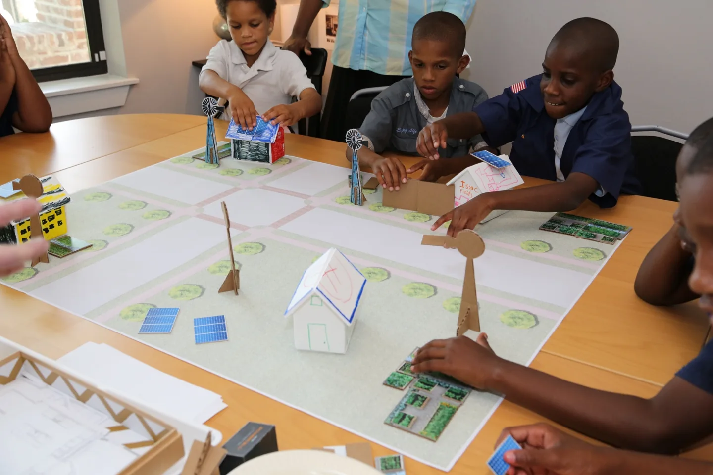 Children assembling a model neighborhood with miniature homes, solar panels, and wind turbines.